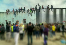 People sitting on a border wall with crowd below.