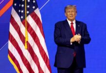 Donald Trump in suit near the American flag on stage.