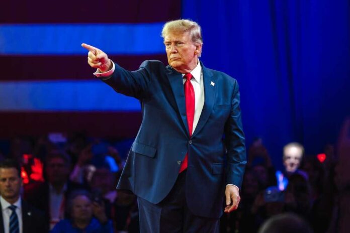 Man in suit pointing on stage with audience behind.