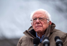 Elderly man in glasses speaks at an outdoor event.