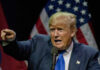 Man pointing while speaking at podium with American flags.
