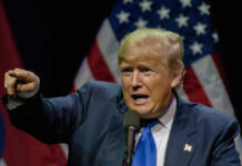 Man pointing while speaking at podium with American flags.