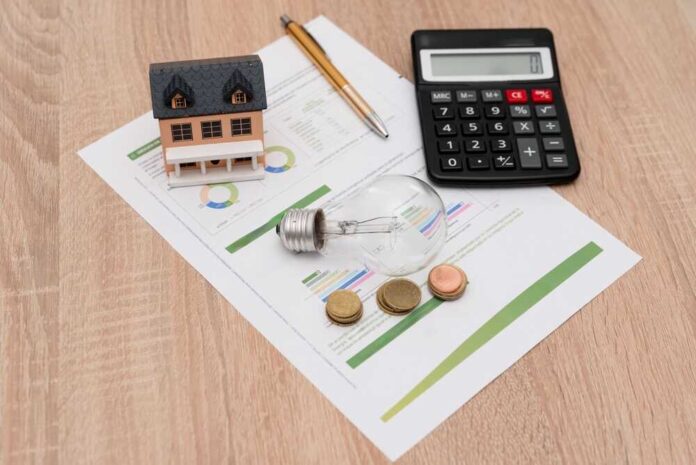 A miniature house model, light bulb, coins, calculator, and financial documents on a wooden surface
