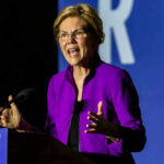 Elizabeth Warren speaking at podium wearing purple jacket.