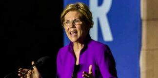 Elizabeth Warren speaking at podium wearing purple jacket.