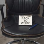 A black office chair with a notepad saying 'BACK TO WORK' and a blue pen on it