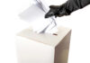 Gloved hand placing multiple papers in a ballot box.