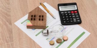 A wooden house model, light bulb, coins, calculator, and financial documents on a wooden surface