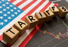 Wooden blocks spelling 'TARIFFS' on a background of the US flag and financial charts