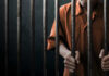 Man in orange prison uniform behind jail bars.