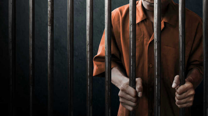 shutterstock_744470836 Man in orange prison uniform behind jail bars.