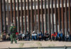 Border patrol officer watches group sitting by a fence.