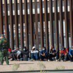Border patrol officer watches group sitting by a fence.