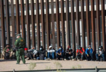 Border patrol officer watches group sitting by a fence.