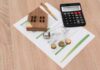 A wooden house model, light bulb, coins, calculator, and financial documents on a wooden surface