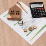A wooden house model, light bulb, coins, calculator, and financial documents on a wooden surface