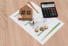 A wooden house model, light bulb, coins, calculator, and financial documents on a wooden surface
