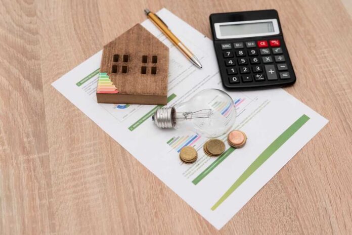 A wooden house model, light bulb, coins, calculator, and financial documents on a wooden surface