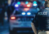 Police officer stands near patrol car with lights flashing.