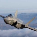 F-35 fighter jet flying above clouds with mountains in the background
