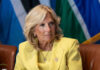 Jill Biden in a yellow blazer sitting, colorful flags behind.