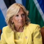 Jill Biden in a yellow blazer sitting, colorful flags behind.
