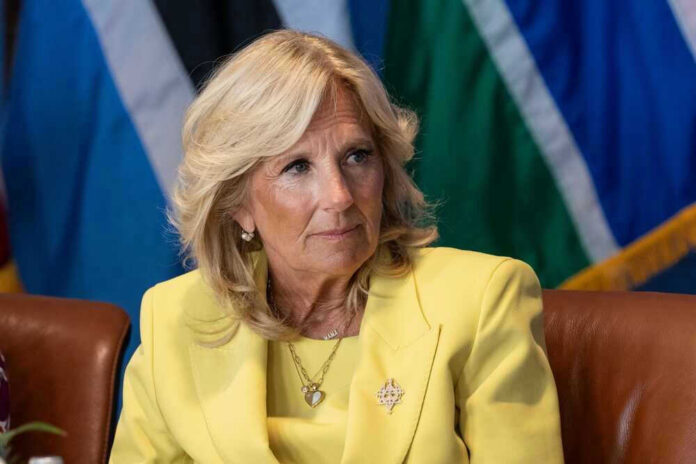 shutterstock_2330920993 Jill Biden in a yellow blazer sitting, colorful flags behind.