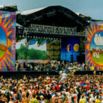 A large crowd at an outdoor music festival with a vibrant stage