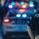 Police officer stands near patrol car with lights flashing.