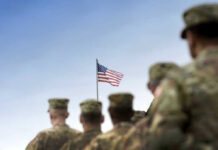 Soldiers in uniform facing American flag outdoors.