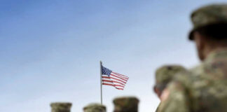 Soldiers in uniform facing American flag outdoors.