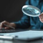 Person using a magnifying glass while working on a laptop