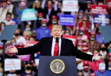Donald Trump speaking at a rally with arms outstretched at a podium