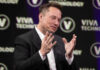 Elon Musk speaking on stage with hands raised during a talk